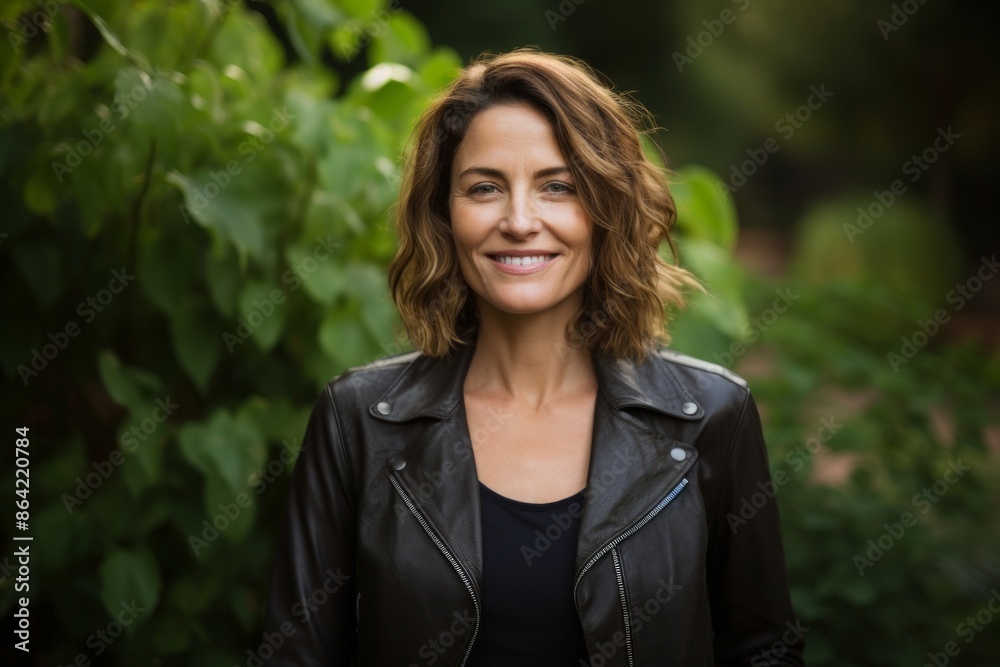 Portrait of a smiling woman in her 40s sporting a stylish leather blazer isolated in lush green garden Portrait of a smiling woman in her 40s sporting a stylish leather blazer isolated in lush green garden