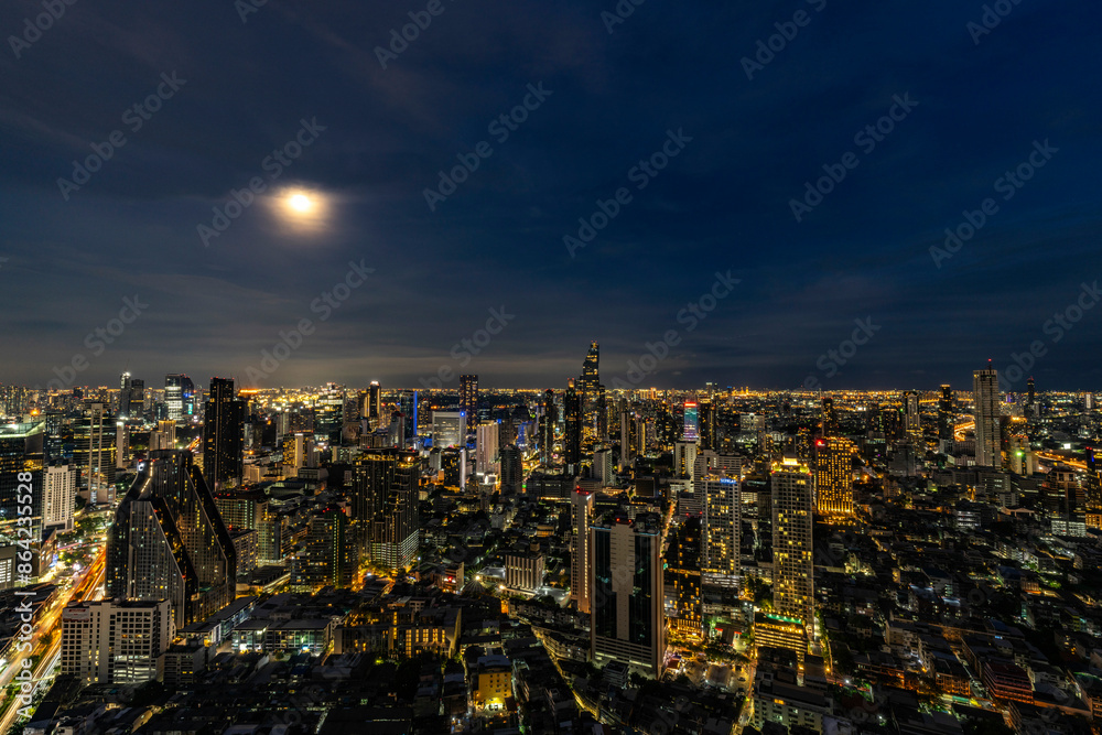 Obraz premium A breathtaking night view of Bangkok's skyline illuminated by city lights and a full moon. The cityscape showcases the blend of modern skyscrapers and vibrant urban life, reflecting the city's dynamic