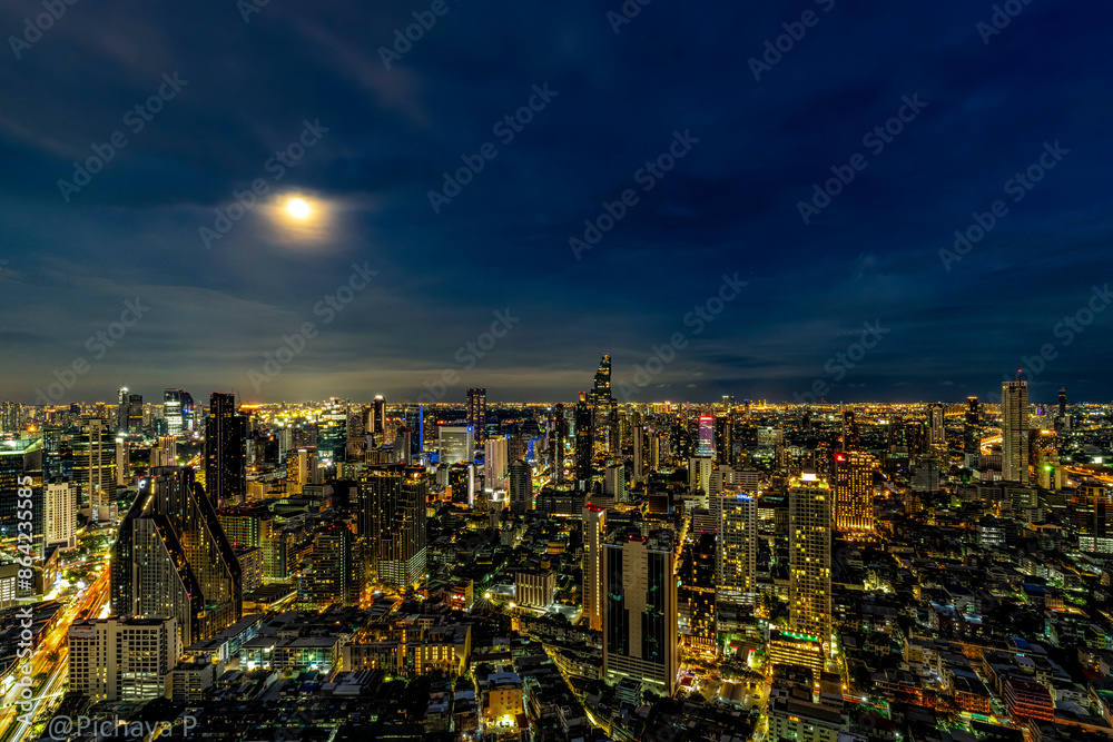 Obraz premium A breathtaking night view of Bangkok's skyline illuminated by city lights and a full moon. The cityscape showcases the blend of modern skyscrapers and vibrant urban life, reflecting the city's dynamic
