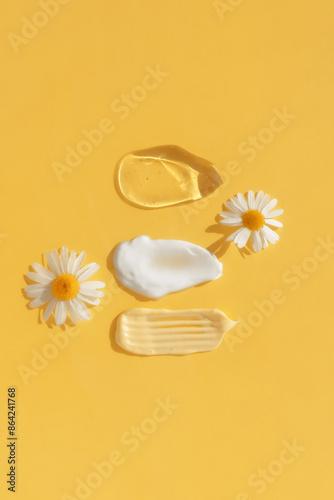 Smears of yellow, white cream and transparent gel or serum with chamomile flowers on a yellow background. The texture of a cosmetic skin care product. The concept of natural cosmetics. Top view.