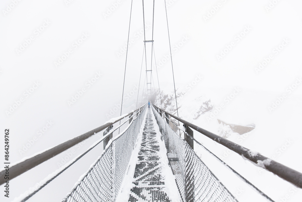 Obraz premium Hängebrücke im Schneesturm. Fußgänger überquert schmale Hängebrücke im Nebel. Suspension bridge in a snowstorm. Pedestrian crosses narrow suspension bridge in the fog.