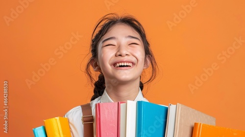 Holding a pile of colorful textbooks, an Asian student in uniform laughs infectiously against a bold orange backdrop. Her genuine happiness and eagerness to learn make her a perfect image for photo