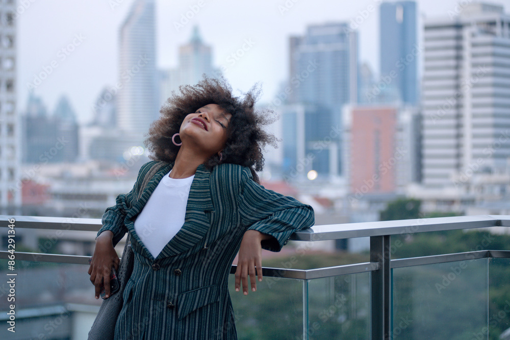 Obraz premium African businesswoman relaxes when she on the rooftop getting some fresh air after a tense meeting, tall building in the background