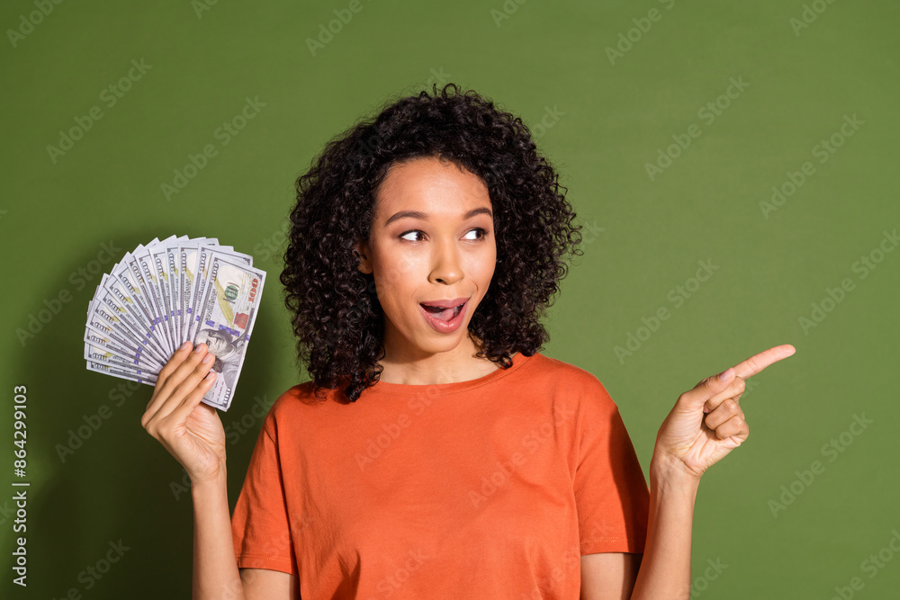 Photo portrait of pretty young girl hold money look point empty space wear trendy orange outfit isolated on khaki color background Photo portrait of pretty young girl hold money look point empty space wear trendy orange outfit isolated on khaki color background