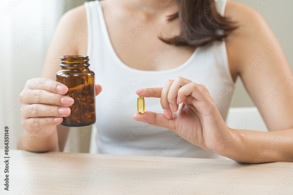 Fototapeta premium Dietary supplement concept, happy asian young woman hand holding fish oil pills, drugs medical tablet pouring capsules from medication bottle, take vitamin treatment for skin, hair at home, healthcare