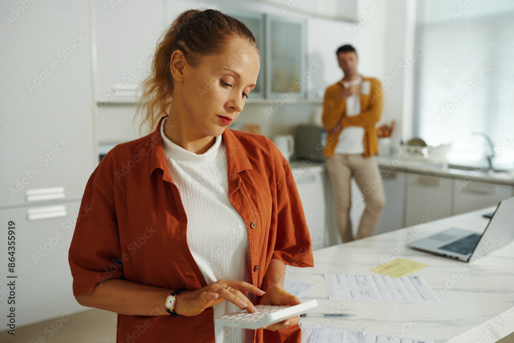 Young woman calculating the bills at home with her husband in background Young woman calculating the bills at home with her husband in background