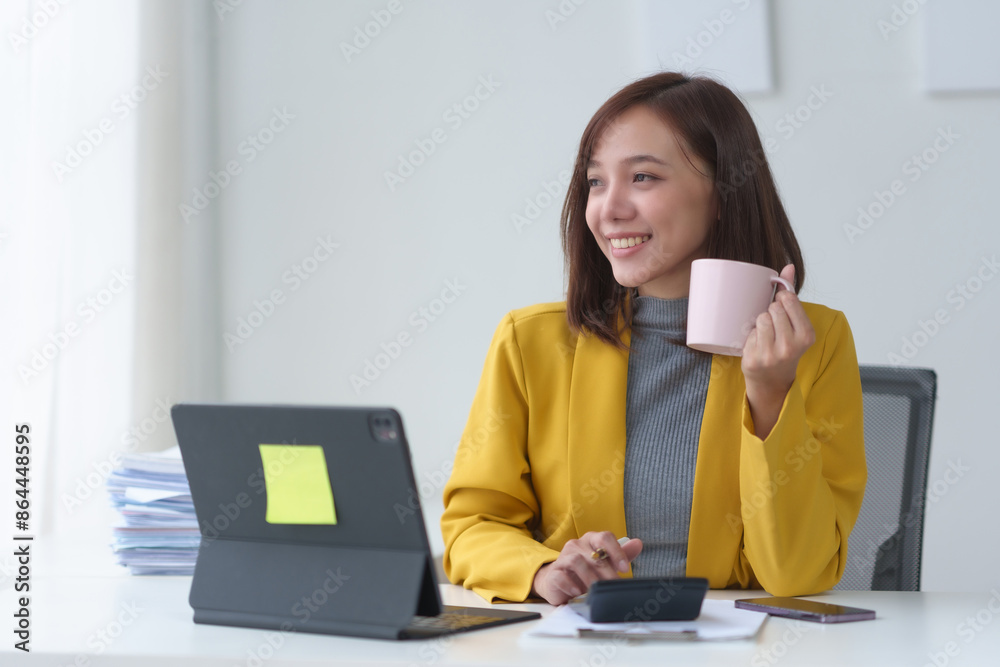 Smiling businesswoman holding coffee cup, working at desk with laptop, papers, and smartphone, wearing yellow jacket in modern office Smiling businesswoman holding coffee cup, working at desk with laptop, papers, and smartphone, wearing yellow jacket in modern office