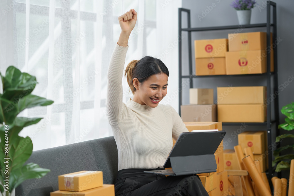 Woman celebrating success while working with a laptop from home. Surrounded by cardboard boxes, she raises her fist in excitement. Woman celebrating success while working with a laptop from home. Surrounded by cardboard boxes, she raises her fist in excitement.