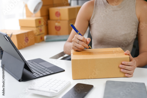 Woman preparing package for shipping, writing address on cardboard box, working with tablet and calculator in home office.