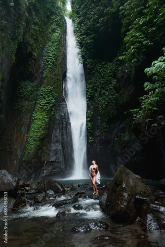 A woman in a black bikini and white shirt stands on rocks at the base of a tall waterfall in a lush, tropical environment.