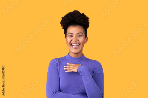 A young woman with an afro smiles broadly while standing against a bright yellow backdrop. She wears a purple turtleneck shirt and has her hands clasped in front of her.