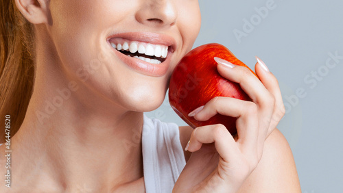A close-up photo of a woman with long blonde hair holding a red apple in her right hand. She is smiling with her mouth open and showing her white teeth, cropped
