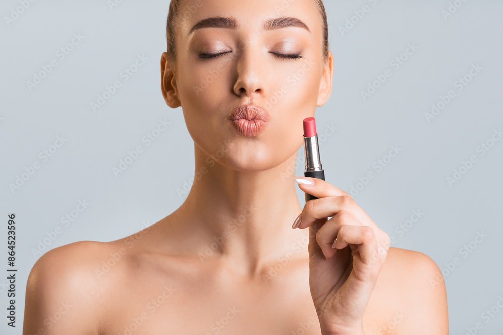 A young woman with closed eyes is holding a pink lipstick in her right hand and applying it to her lips. The woman is wearing no clothing and is looking slightly down. A young woman with closed eyes is holding a pink lipstick in her right hand and applying it to her lips. The woman is wearing no clothing and is looking slightly down.