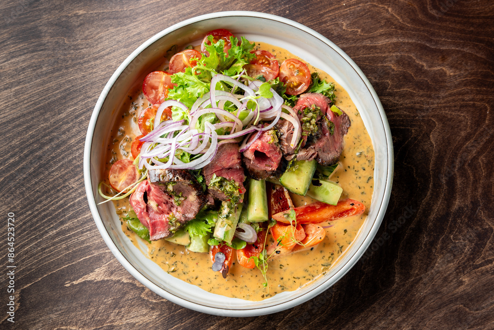 A vibrant salad with medium-rare steak slices, assorted vegetables, and creamy dressing in a white bowl