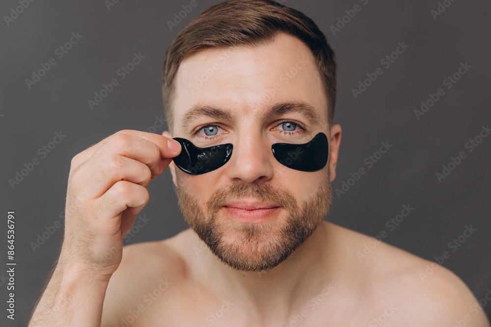 Close-up portrait of handsome bearded man applying black patches under eyes. Attractive guy caring about his appearance on grey background Close-up portrait of handsome bearded man applying black patches under eyes. Attractive guy caring about his appearance on grey background