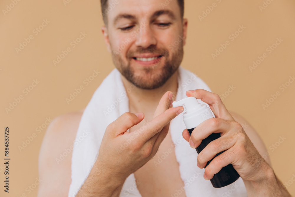 Close-up portrait of cute happy bearded man with towel around neck applying face wash foam on beige background. Beauty and care concept