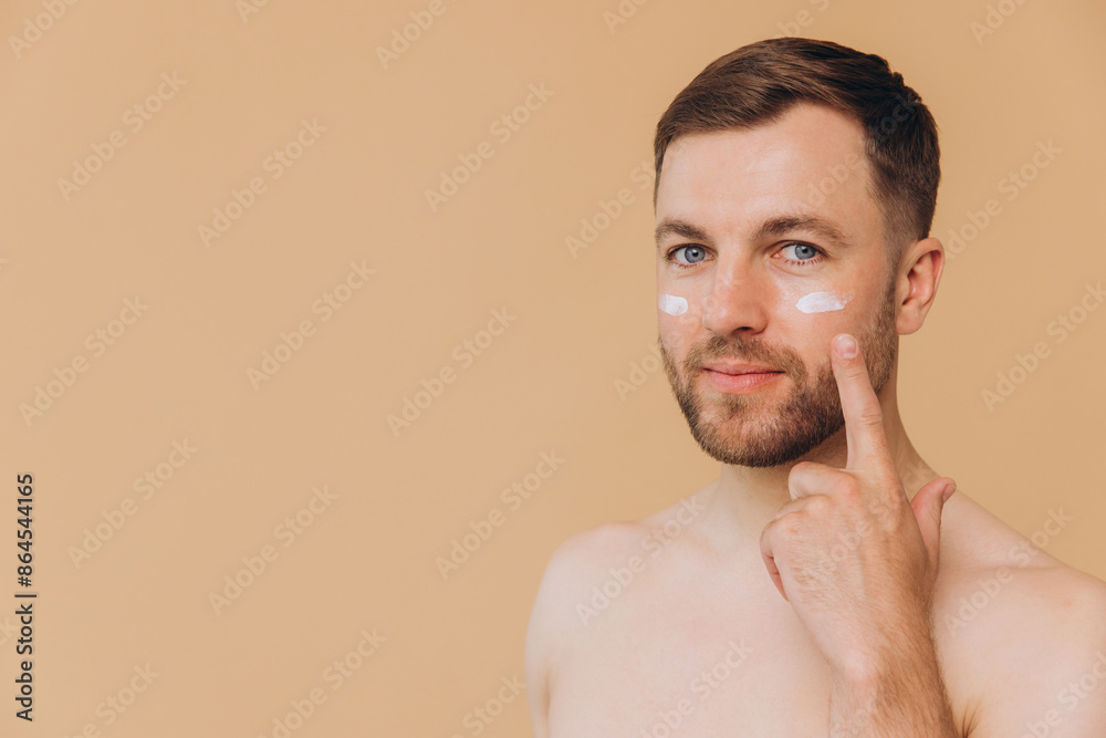 Obraz premium Attractive bearded man with healthy skin, applies cream moisturizer for anti wrinkle or anti aging, cares of body, poses naked or shirtless, isolated over beige background with free space.