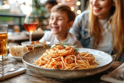 A smiling family is seated around a wooden table outdoors, sharing a meal with plates of spaghetti, drinks, and good times in a warmly lit eating area. photo