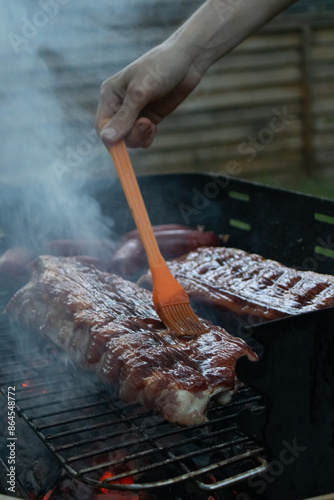 Foto A close-up of a hand basting BBQ ribs with sauce on a grill, surrounded by smoky ambiance