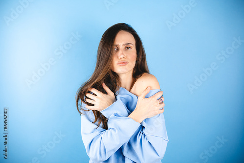 Young woman freezes in long sleeves top warms up by hugging herself, seasonal changes low temperature heating problem, looks at camera, chills feeling of coldness concept, isolated on blue background.
