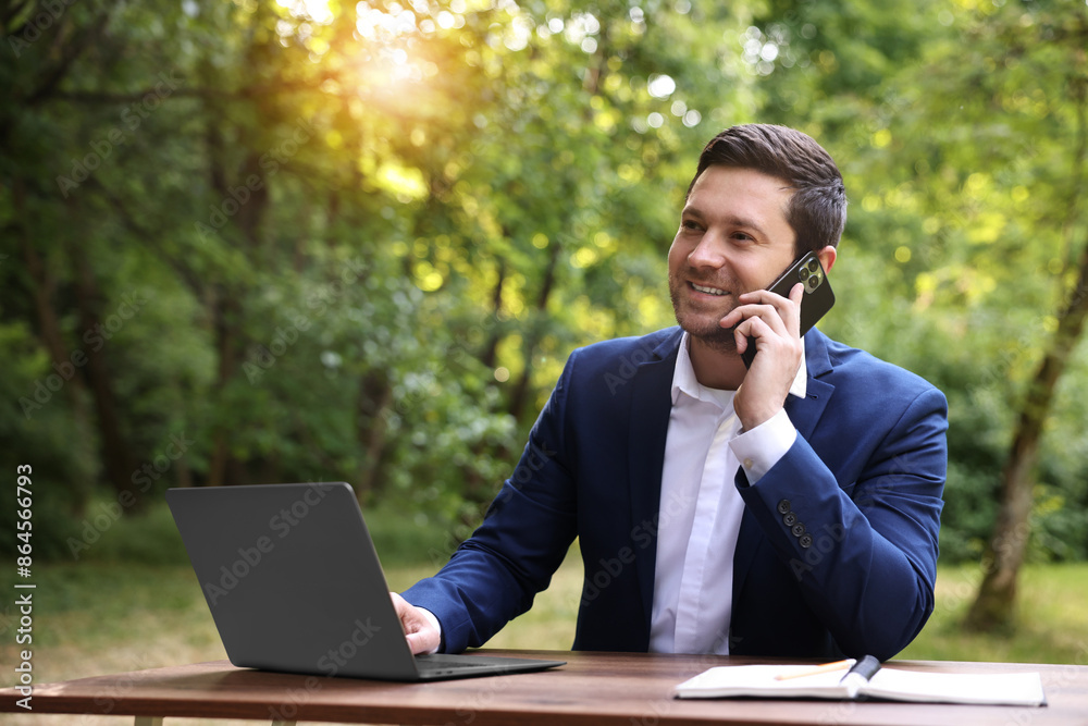 Smiling businessman talking on smartphone at table with laptop outdoors. Remote job Smiling businessman talking on smartphone at table with laptop outdoors. Remote job