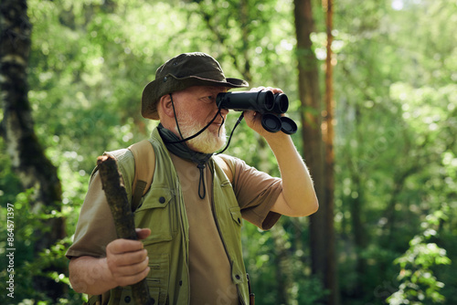 Medium shot of senior Caucasian naturalist looking through binoculars while working in forest on summer day