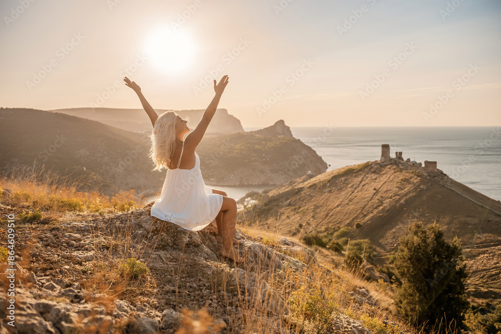 Happy woman is sitting on a hillside, wearing a white dress. She is surrounded by a beautiful landscape, with a body of sea in the background. Concept of peace and happiness. Happy woman is sitting on a hillside, wearing a white dress. She is surrounded by a beautiful landscape, with a body of sea in the background. Concept of peace and happiness.
