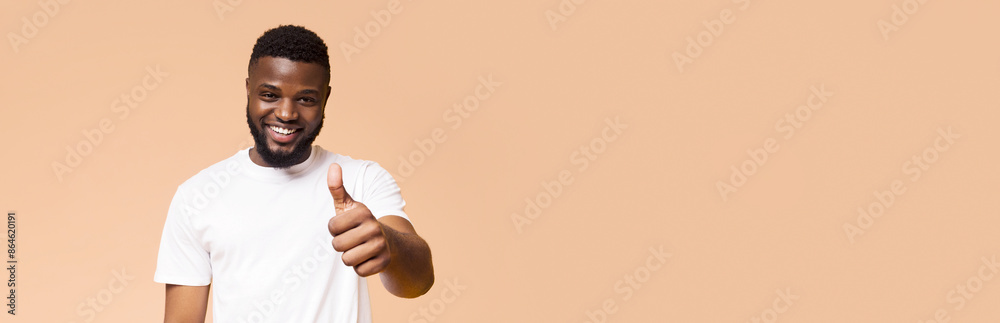 A smiling black man with short, dark hair and a beard is wearing a white T-shirt. He is giving a thumbs up gesture with his right hand. The background is a light peach color, copy space A smiling black man with short, dark hair and a beard is wearing a white T-shirt. He is giving a thumbs up gesture with his right hand. The background is a light peach color, copy space