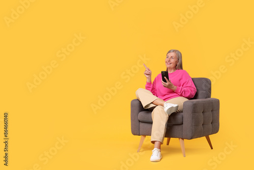 Senior woman with phone on armchair against orange background, space for text