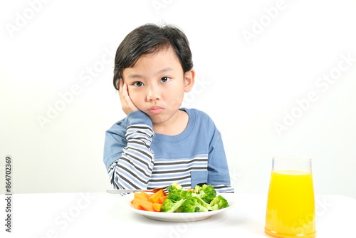 A young boy in a striped blue shirt looks bored while sitting at a table with plate of vegetables and a glass of orange juice.Perfect for themes on children's nutrition, picky eating,and healthy diets