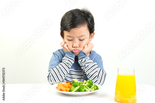Canvas Print A young boy in a striped shirt sits at a table,looking unhappy with his plate of vegetables and a glass of orange juice