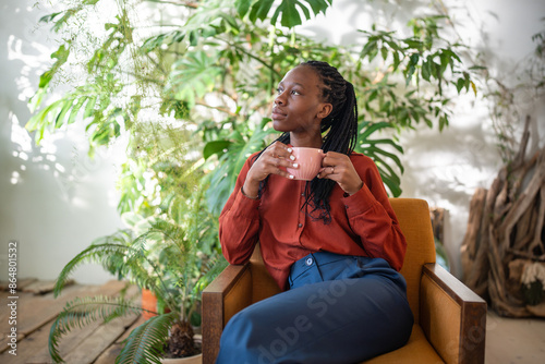 Dreaming african american woman holding cup of coffee sitting on chair in urban jungle interior looking aside window, thinking. Thoughtful black female resting on armchair surround houseplants.  photo