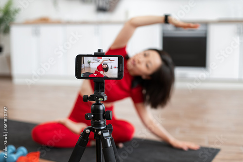 Woman in red outfit filming online yoga class at home using smartphone. Fitness enthusiast recording video content for health and wellness vlog. Bright kitchen backdrop indicates domestic setting.