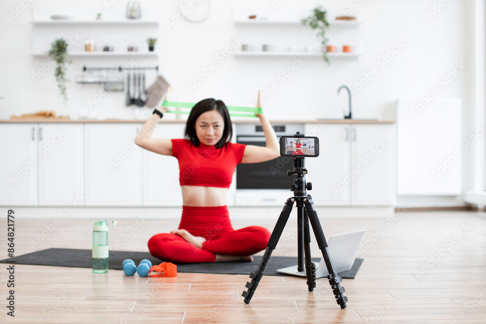 Fototapeta premium Focused woman in red activewear filming fitness tutorial at home using resistance bands. Exercise equipment, water bottle, and laptop nearby, creating realistic workout setting