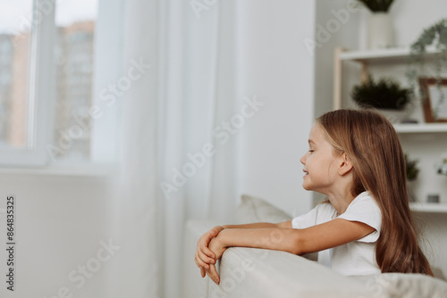 Free Download Curious little girl on a cozy couch gazes out the window, captivated by something in the distance - GFXLoad