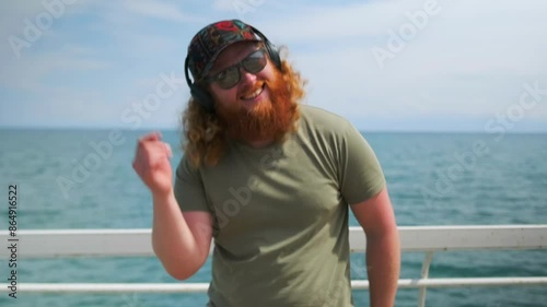 Young caucasian redhead man dances in sunglasses on wooden pier on seashore, positive hipster celebrating holiday. Mid shot.