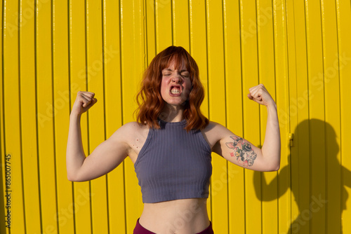 Redhead woman wearing purple crop top flexing muscles in front of yellow wall on sunny day photo