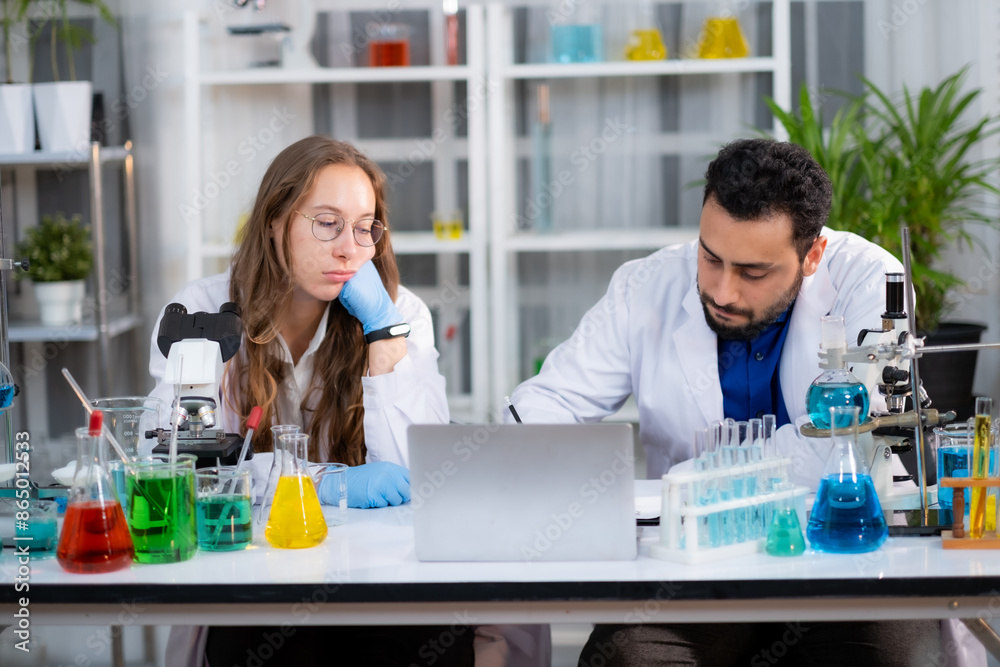Young White Scienctist woman sitting at experiment work desk with boring emotion, while her friend recording experiment result in Lab room. Young White Scienctist woman sitting at experiment work desk with boring emotion, while her friend recording experiment result in Lab room.