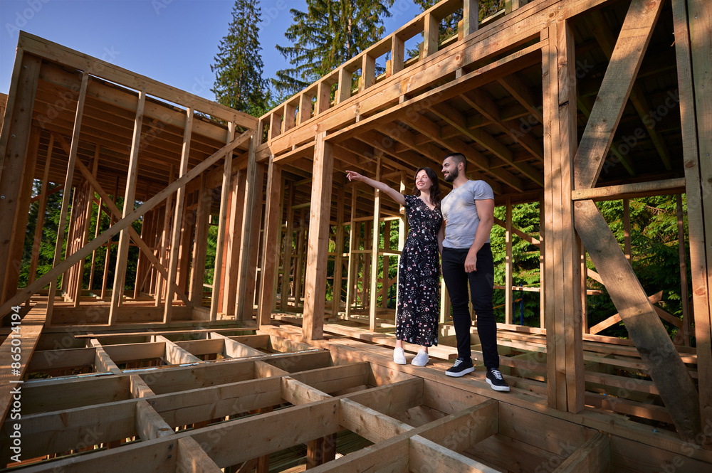 Man and woman examining their future wooden frame dwelling nestled near the forest. Youthful couple at construction site in early morning. Concept of contemporary ecological construction. Man and woman examining their future wooden frame dwelling nestled near the forest. Youthful couple at construction site in early morning. Concept of contemporary ecological construction.