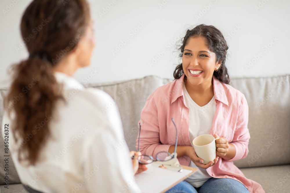 A woman wearing a pink shirt sits on a couch and smiles while holding a cup of coffee. She is talking to another woman sitting next to her, who is wearing a white shirt. A woman wearing a pink shirt sits on a couch and smiles while holding a cup of coffee. She is talking to another woman sitting next to her, who is wearing a white shirt.