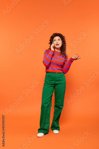 A girl with curly hair wears a striped sweater and green pants against an orange background, talking on phone and looking up.