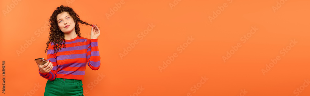 A young woman with curly hair poses against a bright orange background, holding a phone and twirling a lock of her hair. A young woman with curly hair poses against a bright orange background, holding a phone and twirling a lock of her hair.