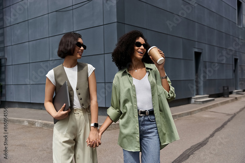 Two women walk hand-in-hand, smiling and enjoying a sunny day in the city.