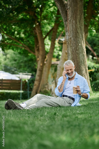 A mature gay man with tattoos and a grey beard sits on the grass outdoors. He is on a phone call and enjoying a coffee.
