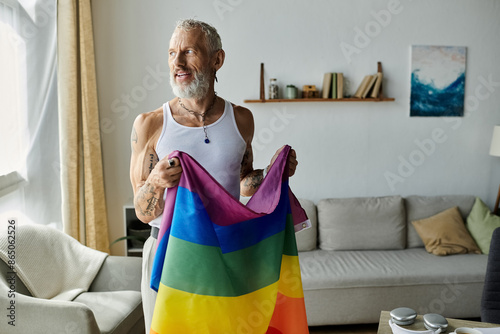 фотографія A mature gay man with tattoos and grey hair smiles and holds a pride rainbow flag in his living room