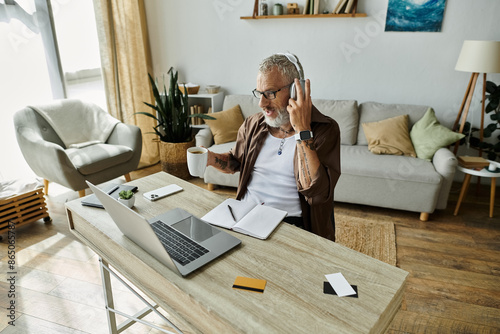 A tattooed man with grey hair works on a laptop at a desk, wearing headphones and holding a phone and coffee cup while working remotely. © Bliss