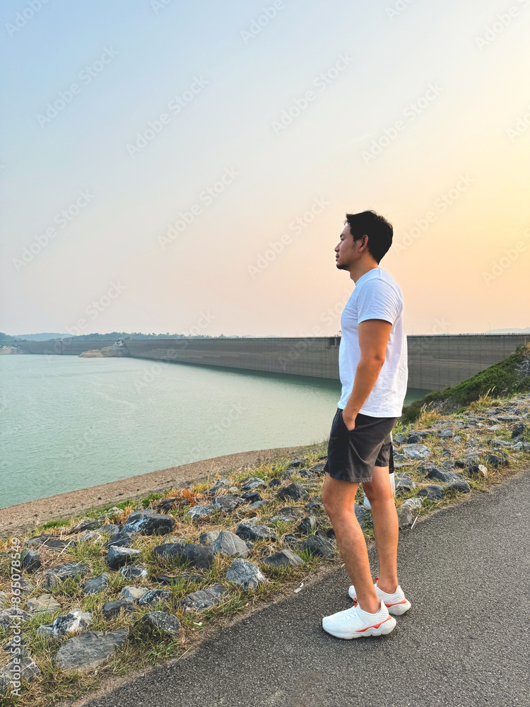 Portrait of asian Thai man standing with dam view and sunset sky, happy traveling concept.