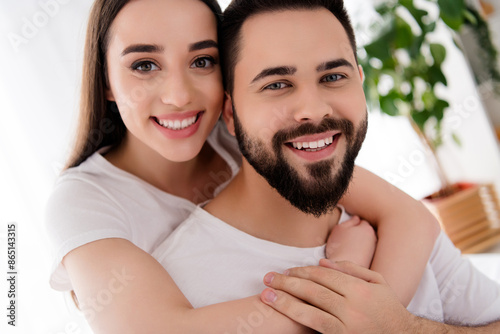 Photo of toothy beaming couple wear white t-shirts embracing indoors apartment bedroom