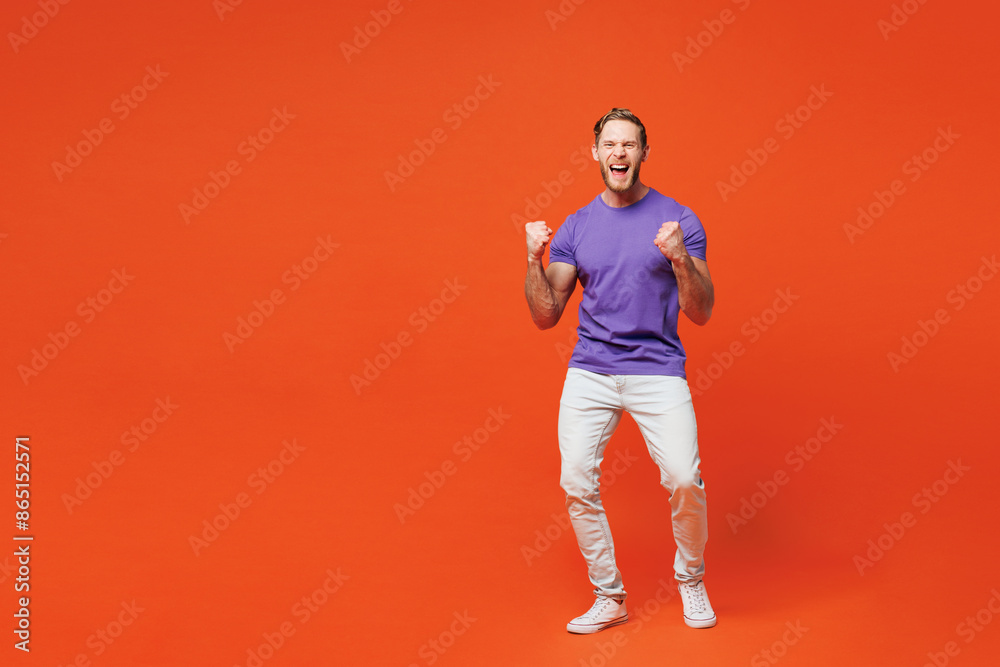 Full body young smiling happy man he wearing purple t-shirt casual clothes doing winner gesture celebrate clenching fists say yes isolated on red orange background studio portrait. Lifestyle concept. Full body young smiling happy man he wearing purple t-shirt casual clothes doing winner gesture celebrate clenching fists say yes isolated on red orange background studio portrait. Lifestyle concept.