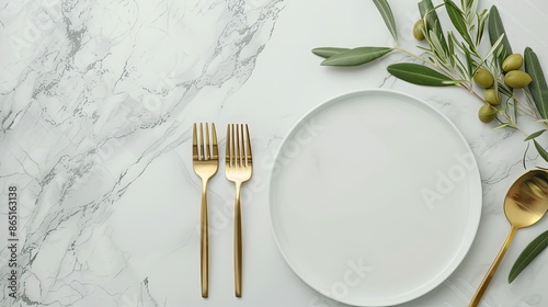 A white marble tabletop with a white plate, gold fork, gold knife, and gold spoon. An olive branch with green leaves extends from the left side of the frame photo